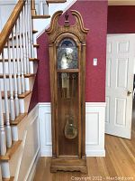 Front view of Howard Miller grandfather clock showing clock face, pendulum, and weights behind glass door, positioned next to staircase.