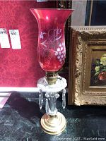 Single lamp with red etched grapevine glass shade, clear glass base with hanging prisms, brass base, sitting on black surface in front of red wallpaper and floral painting.