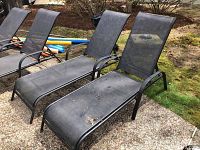 Two black outdoor lounge chairs placed outside on a patio with a gravel surface and grass nearby. Both chairs have mesh seating fabric stretched over black metal frames with armrests and slanted backs for reclining.
