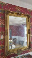 Frontal photo of the large rectangular mirror with ornate gold frame hanging on a wall with a floral red wallpaper.