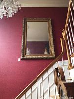 Front view of the large rectangular mirror with ornate gold frame mounted on a red wall by a staircase, showing reflection of a chandelier and parts of the room.