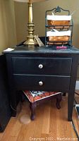 Side view of one black nightstand showing two drawers with silver knobs, lamp, and basket organizer on top