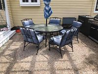 Five metal patio chairs with blue seat and back cushions arranged outdoors around a glass-top patio table with umbrella.