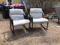 Front view of two metal framed patio rocking chairs with gray/white patterned cushions on a textured patio surface.