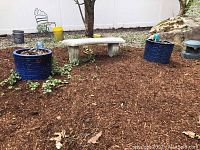 Wide shot of the two matching blue ceramic planters placed outdoors on mulch ground next to a concrete bench and some greenery.