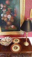 Decorative painted glass bowl, decorative plates, and gold-toned lamp with black lampshade on wooden surface with floral painting in background.