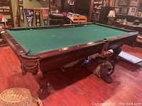 Side angled view of the pool table showing the claw feet, green felt surface, and braided tassel pockets, with partial view of surrounding basement area.