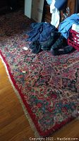 Photo of the red and blue colored Oriental rug laid on a hardwood floor, with clothing and boots placed on top. Shows pattern detail and condition.