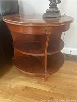 Side view of the round wooden side table showing two curved shelves, vertical supports with carved medallions, and rounded feet on hardwood floor.