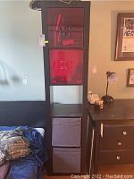 Front view of the black five shelf storage unit showing the two red translucent doors and fabric drawer, placed beside a bed and nightstand.