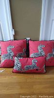 Three matching decorative throw pillows with zebra designs in black and white on red fabric with black piping. Two large square pillows and one smaller rectangular pillow arranged on wood surface against a wall.