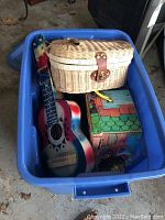 Blue plastic bin containing a woven doll picnic basket, colorful toy guitar and decorative puzzle box.