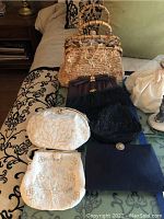 Seven vintage handbags assembled on a bed, showing various styles including white and black beaded evening bags, a straw handbag, blue and black clutches