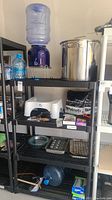 Photo of shelving containing a ceramic water dispenser with water jug, blue water jug, baking pans, squatty potty, survival food kits, small white appliance, and boxes of kitchen items.