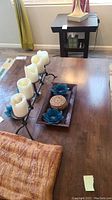 Close-up photo of black metal candelabra with five white pillar candles and wooden tray with lotus candle holders and coasters, with warm-toned throw partially visible.