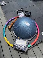 Blue half balance ball with textured grip alongside wooden MA roller and colorful segmented hula hoop on black floor mats, also showing workout manuals