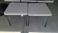 Two beige plastic folding tables side by side, showing tabletops and legs, tags visible on one table