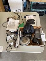Photo of a cardboard box containing various small electronics including cords, power strip, black Bose pouch, and a digital clock or timer.