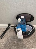 Sharper Image ultrasonic jewelry cleaning device with transparent lid opened. Two partial bottles of blue Isonic Jewelry Cleaner Concentrate inside along with attached power cord placed on the floor.