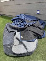 Multiple moving blankets piled on grass, showing blue and gray quilted fabric