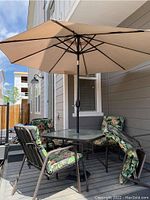 Full view of patio dining set showing glass table, four chairs with floral cushions, large beige umbrella and weighted black stand on deck.