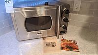 Stainless steel Wolfgang Puck pressure oven with three control knobs and a viewing window on the door, placed on a kitchen counter with manual and recipe booklet in front.