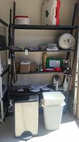 Full view of garage shelving showing black and white trash bins, mailers, small boxes, and various garage items like staple guns and packing tape.