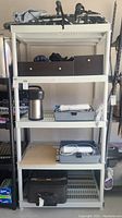 Full view of white plastic shelving unit with several items including thermos, electronic parts, bins, and suitcase.