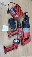 Photo showing Milwaukee battery charger, battery pack, cordless drill, cordless reciprocating saw, and cordless handheld work light all on wood surface.