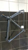 Top view of matte black carbon fibre bike frame with attached pedal on a tiled floor, showing main triangle and rear triangle structure.