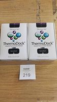 Photo showing both sealed Medisana ThermoDock infrared thermometer module boxes side by side on a wooden surface with a lot number card.