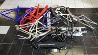 Assorted colored metal bicycle frames including blue, red, white, purple, and black e-bike frames arranged on a tiled floor.