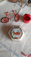 Photo of red decorative metal bicycle, hexagonal porcelain music box with Christmas sleigh artwork, and small red porcelain basket on a light fabric surface.