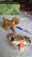 Photo showing both the light brown glass horse with repaired rear leg and the white wooden horse with detailed leather saddle, placed on a white cloth with a pen for scale.