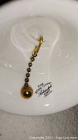 Close-up photo of the underside of the bone china bell showing gold trim, chain with gold beads, and gold spherical weight. Stamped with "Fine Bone China" and brand information.