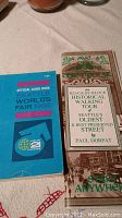 Two books displayed on a light-colored fabric surface; left book is Official Guide Book Seattle World's Fair 1962 with a blue cover, right book is The Block-by-Block Historical Walking Tour with a vintage styled cover.