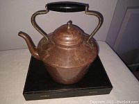 Full side view of copper tea kettle on a black box, showing overall shape, spout, and handle.