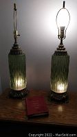 Two identical green glass and brass lamps without shades, both lit showing light from the base and top bulb.