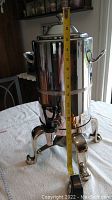 Front view of stainless silver plated coffee urn on metal stand with measuring tape showing height approximately 15 inches