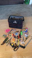 Overall view of Husky tool bag with assorted hand tools and accessories laid out on floor in front. Tools include pliers, screwdrivers, hammer, utility knife, tape, WD-40 spray, string and small hardware items.