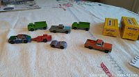 Wide shot of 7 old metal toy cars displayed on a white cloth surface, including 2 green shuttle trucks, silver and orange pickups, a purple car, a red car with trailer, and a multi-color race car. Two yellow Matchbox boxes shown to the side.