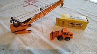 Two photos showing orange Matchbox toys of a Hitachi hydraulic crane and an MB 8 earth mover construction vehicle, each with original yellow Matchbox boxes.