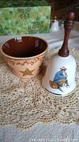 Photo showing Norman Rockwell collectible bell with wooden handle, Star Country ceramic dish with star design, and one crochet doily underneath.