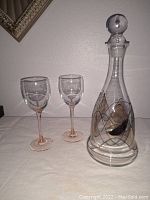 The glass decanter with a glass ball stopper and two wine glasses with decorative patterns on a flat surface.
