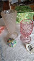 Photo showing the frosted pink glass bubble vase, pink crystal stemmed glass, colorful metal Easter egg, and miniature poodle figurine laid out on a table with a green and gold decorative box in the background.