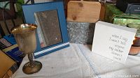 Silver goblet engraved 'Ted', wooden cutting board, organizer box made of wood and metal, and white decorative plaques visible on a cloth surface.