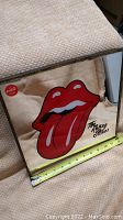 Close-up of lower left corner of mirror showing the red Rolling Stones tongue logo, band signature, and part of the measuring tape for size reference, also showing metal frame and mirrored edge.