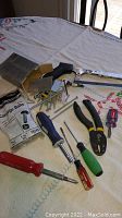 View of various hand tools including multiple screwdrivers, pliers, saw bit driver, mixed screws in an open box, and unopened bag of carriage bolts on a table with tablecloth.