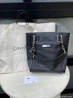 Full front view of black Calvin Klein handbag pictured with original white Calvin Klein shopping bag and receipt tags in front.