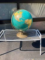 Image of a 16-inch diameter large political world globe showing North and South America, mounted on a bronze-colored metal stand with semi circular meridian, placed on a white metal mesh table.